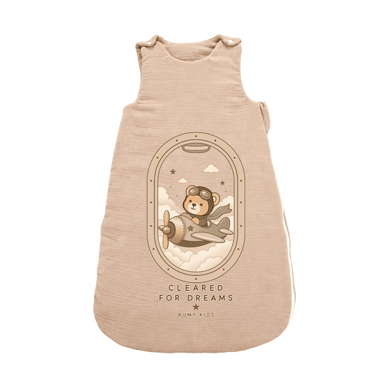 A sleeveless baby sleeping bag in a light beige heathered fabric, featuring a central graphic of a teddy bear pilot flying in a vintage airplane from an airplane window view. Below the illustration, the text reads 'CLEARED FOR DREAMS' and 'KUMPI KIDS'.