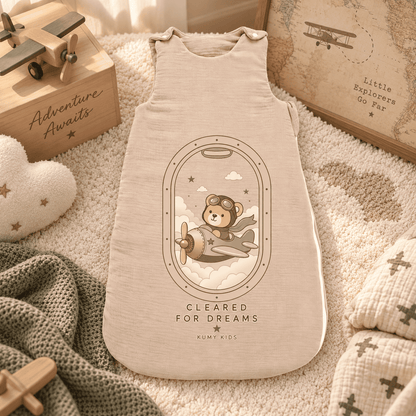 A cream-colored baby sleep sack with a teddy bear airplane graphic, styled on a cozy beige blanket with wooden toys and a knit blanket.