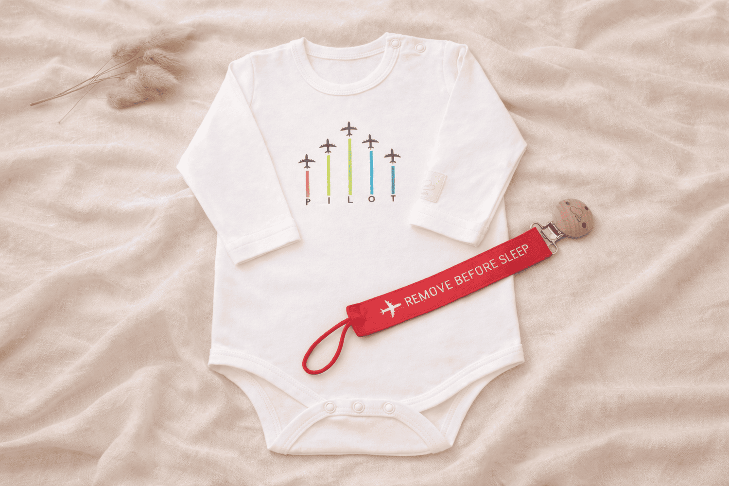 White baby onesie with colorful airplane design and a red 'Remove Before Sleep' strap on a beige surface.