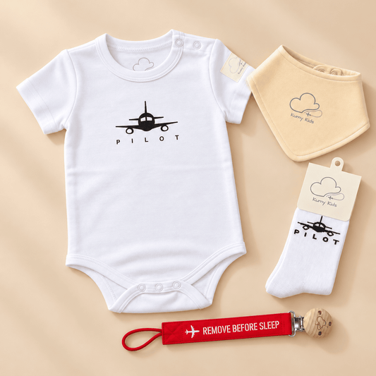 A baby outfit set laid flat on a warm beige background, including a white short-sleeve bodysuit with a black airplane and the word 'PILOT,' a cream bib with a cloud and 'Every Kid,' a pair of white socks with the same airplane graphic, and a red sleep tag that reads 'REMOVE BEFORE SLEEP.'.